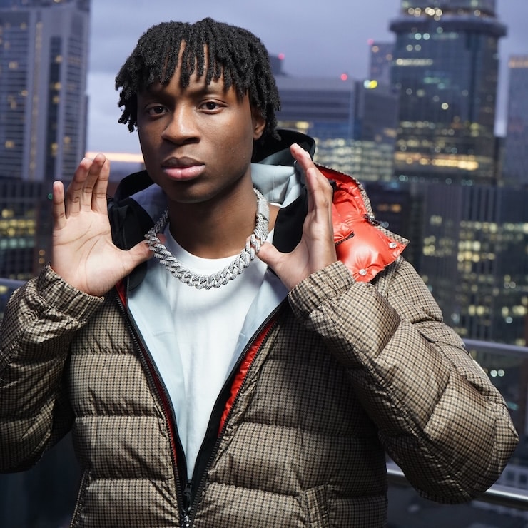 Press shot of rapper Tkandz holding his chain and posing in front of a cityscape